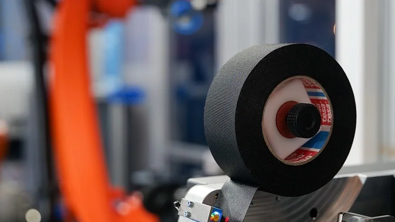 Nahaufnahme eines industriellen Maschinenteils mit einer Rolle Tesaband im Vordergrund und einem orangefarbenen Roboterarm im Hintergrund.