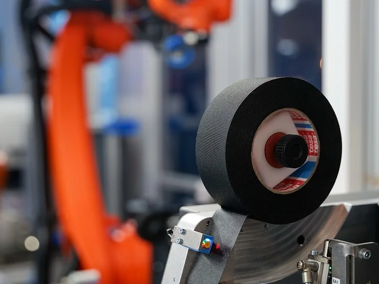 Nahaufnahme eines industriellen Maschinenteils mit einer Rolle Tesaband im Vordergrund und einem orangefarbenen Roboterarm im Hintergrund. (This text has been generated by AI)