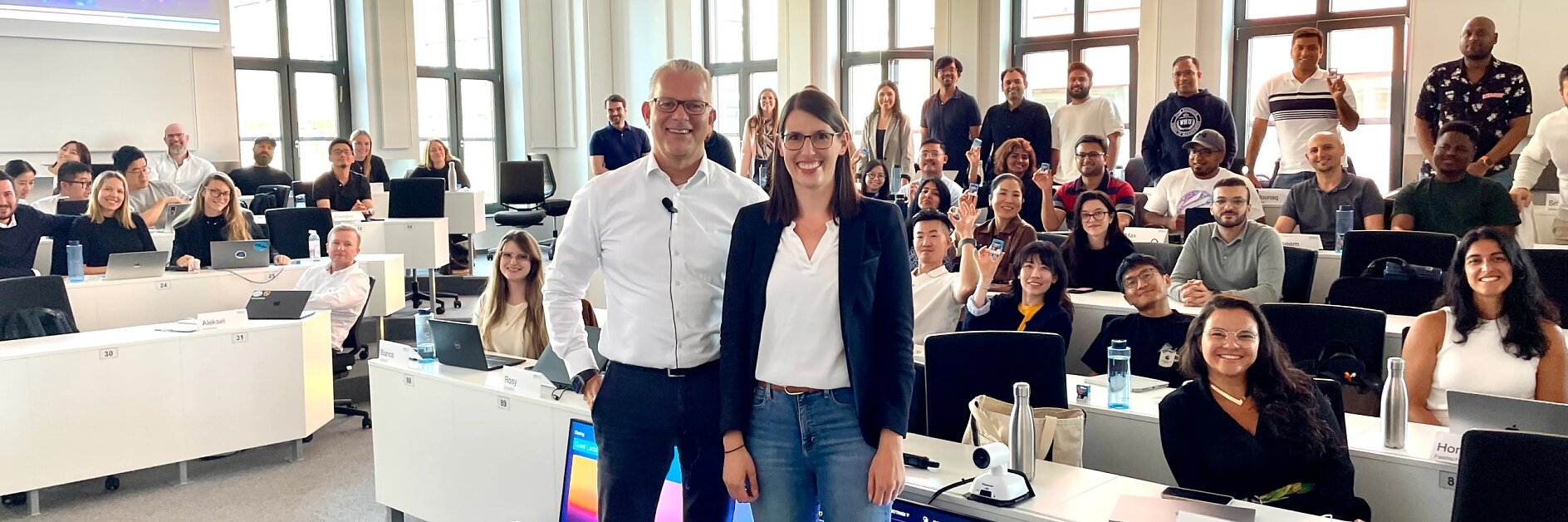 A group poses in a modern classroom with large windows; two in front smile. Desks hold laptops, name tags, and tesa tape rolls. (This text has been generated by AI)