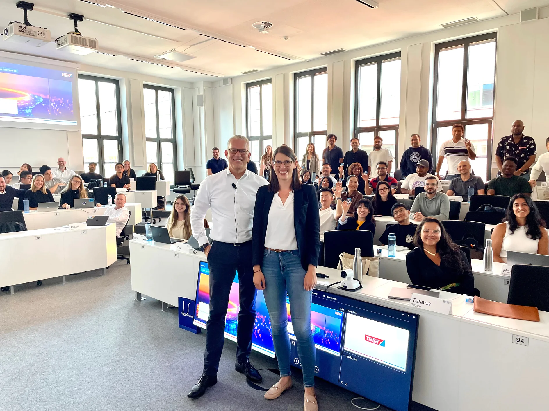 A group poses in a modern classroom with large windows; two in front smile. Desks hold laptops, name tags, and tesa tape rolls. (This text has been generated by AI)
