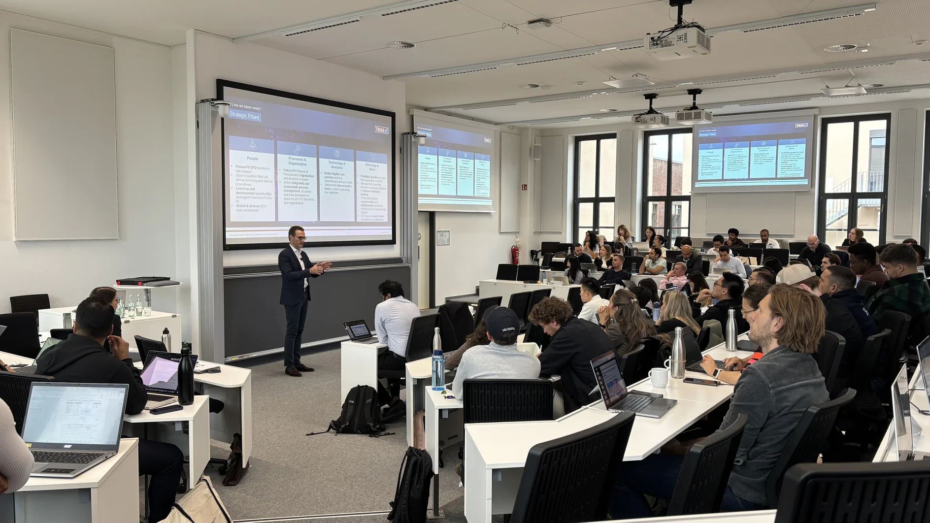 A professor in a suit lectures students with laptops. Two screens show slides. The bright class has tesa tape on the white walls. (This text has been generated by AI)