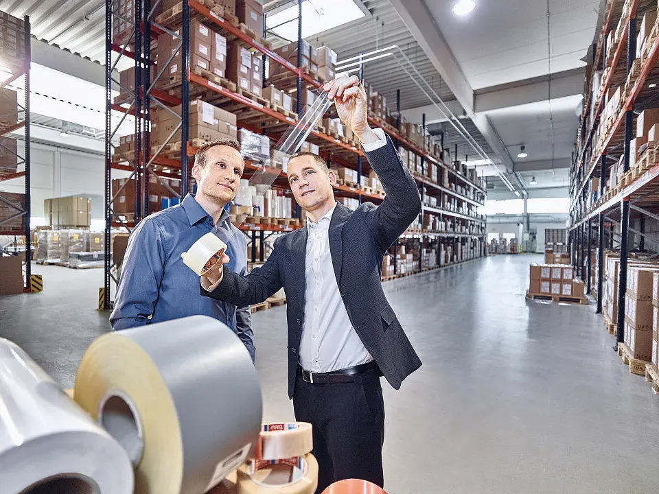 two men in business attire inspecting packaged products in a large, well-lit warehouse with tall shelves filled with inventory tesa (This text has been generated by AI)