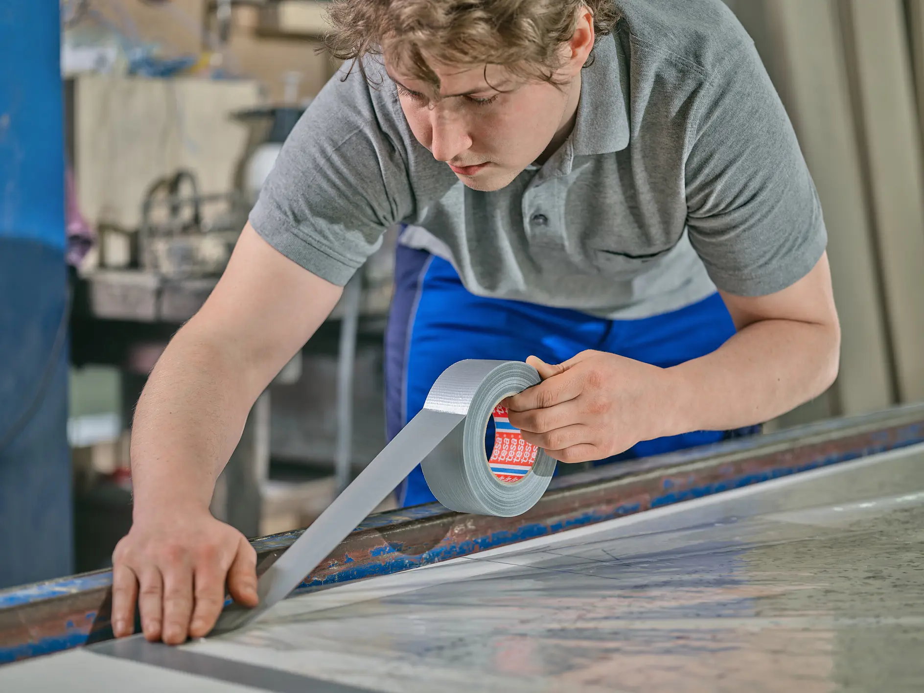 tesa worker in gray shirt and blue pants applying silver duct tape to a large flat surface in an indoor workshop setting (This text has been generated by AI)