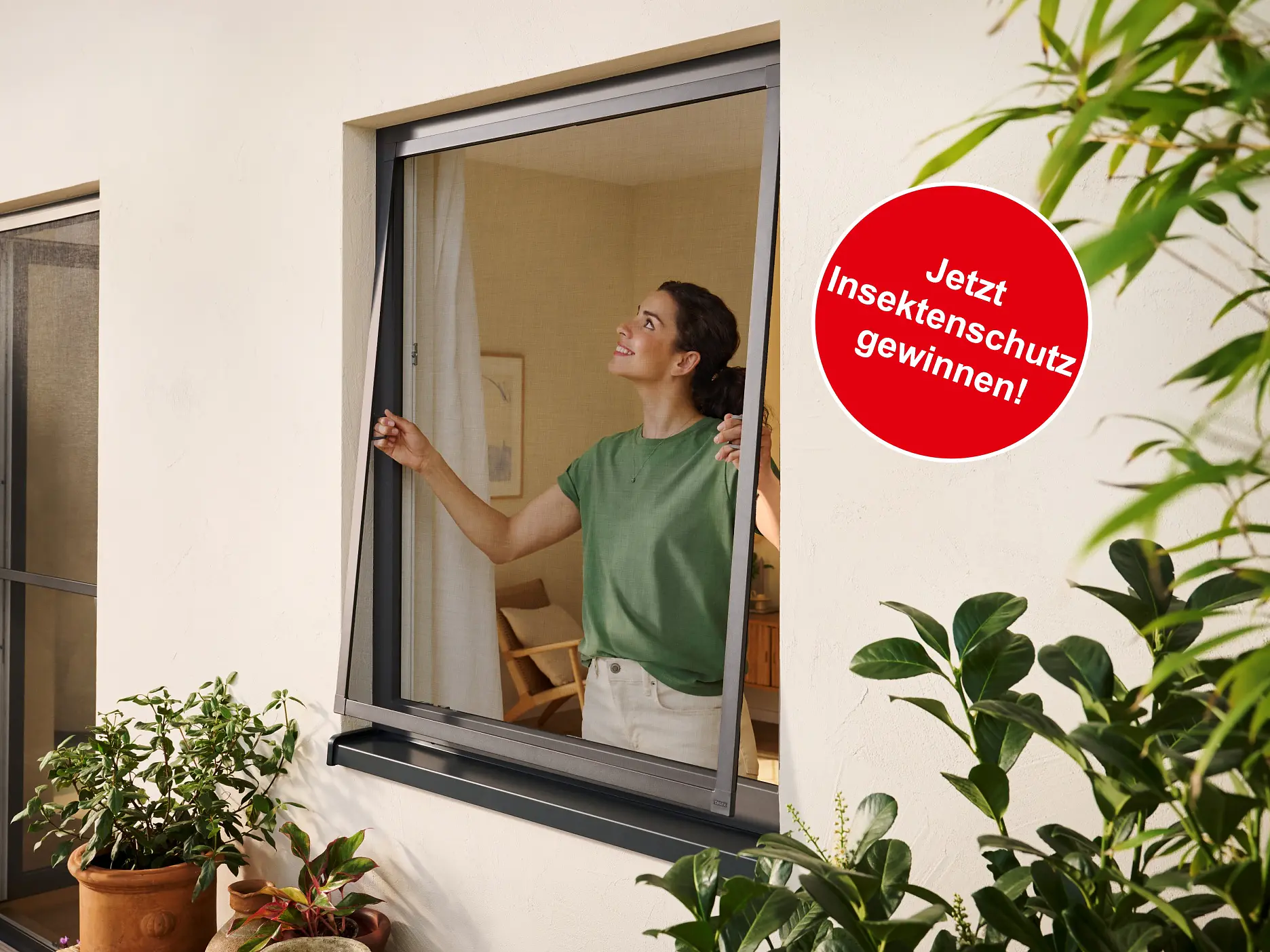 Eine Frau in einem grünen Hemd bringt in einem Raum mit Pflanzen einen Insektenschutz mit Tesafilm an einem Fenster an. Auf einem roten Schild steht 