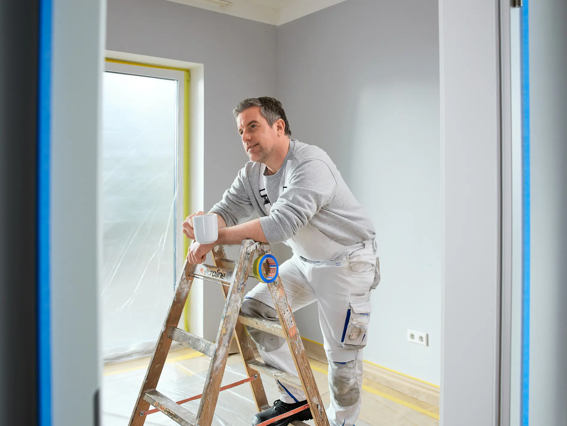 Ein Mann in Malerkleidung steht auf einer Holzleiter, hält eine weiße Tasse und macht eine Pause in einem Raum, in dem tesa-Klebeband an den Leisten zu sehen ist. (This text has been generated by AI)