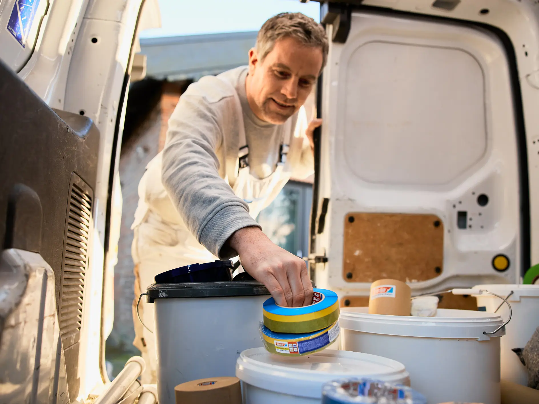Ein Mann in weißem Overall greift nach Rollen tesa-Klebeband in einem Transporter mit Farbeimern und Malerutensilien. Die Türen des Transporters stehen offen und Tageslicht fällt herein. (This text has been generated by AI)