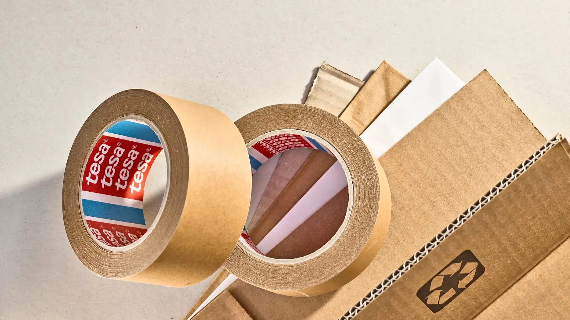 Zwei Rollen braunes tesa Klebeband liegen neben Wellpappe und weißem Papier auf einer hellen Fläche, auf den Etiketten ist tesa zu lesen. (This text has been generated by AI)