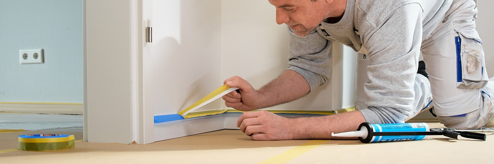 Ein Mann in Arbeitskleidung kniet und bringt tesa Klebeband an einem weißen Türrahmen an; Malerwerkzeuge und tesa Klebeband liegen in der Nähe auf dem Boden.