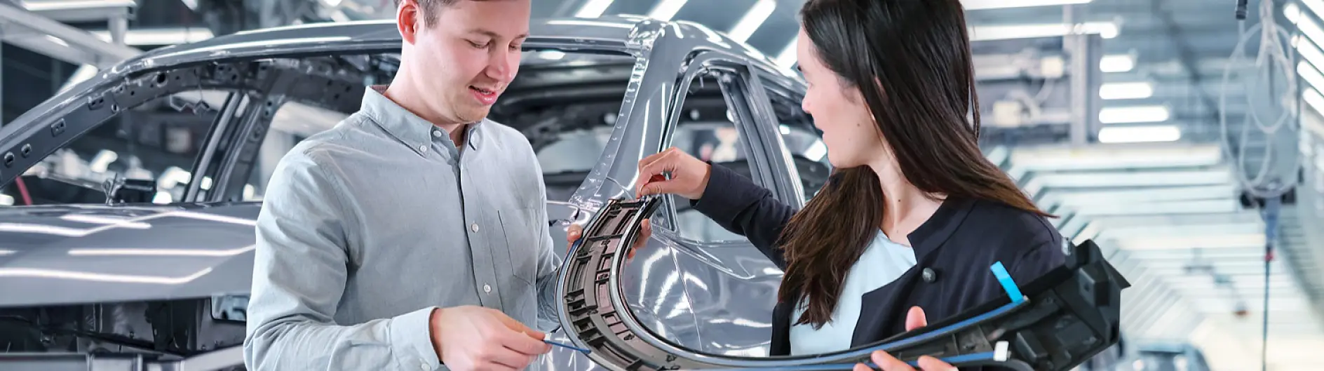 Two people in a factory examine a curved car part, possibly using tesa tape, with assembly equipment and a metallic car frame behind them.