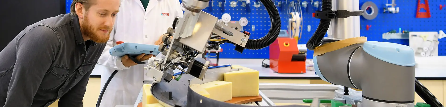 Two people observe a robotic arm using tesa tape on foam materials at a workbench, with tools on a blue pegboard in the background.