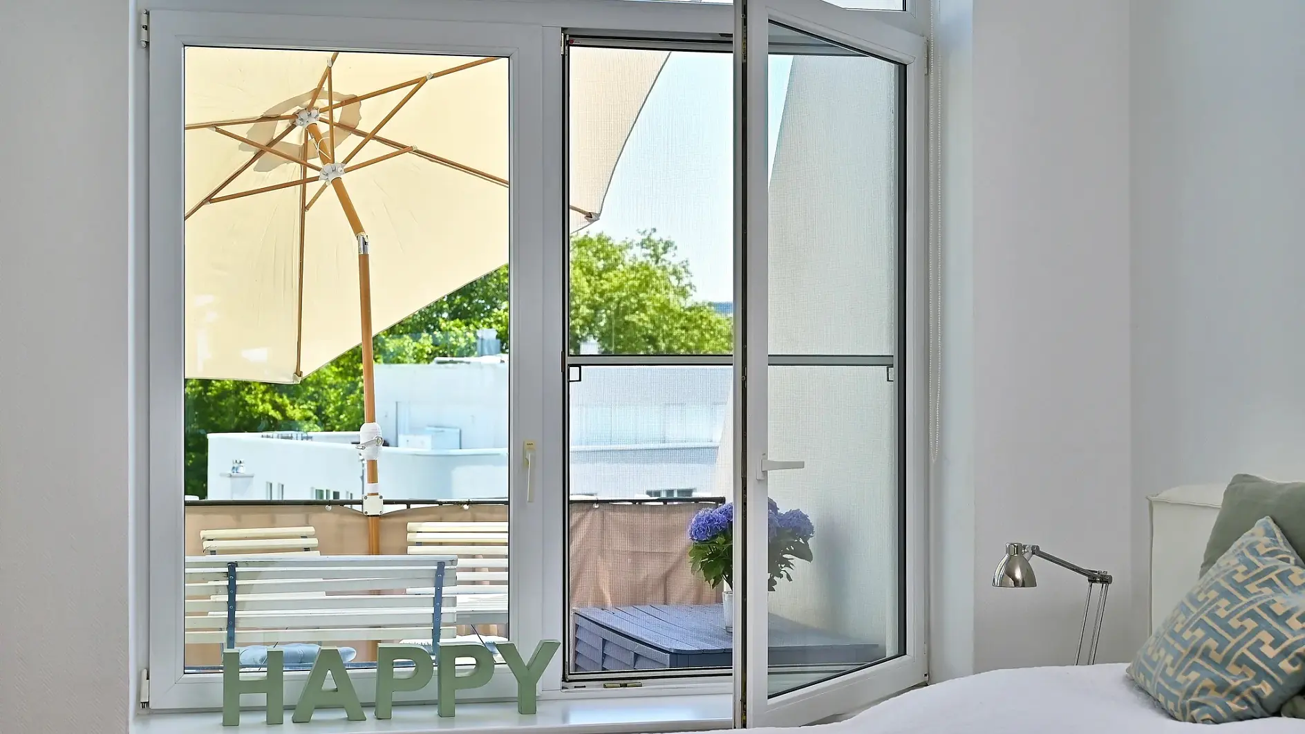 Ein Schlafzimmer mit einem weißen Bett, einer kleinen Lampe und einem Fenster, das auf einen Balkon hinausgeht. Auf dem Balkon stehen ein großer beiger Sonnenschirm, grüne Pflanzen und eine Bank. Auf der Fensterbank stehen grüne Buchstaben, die das Wort „HAPPY“ bilden und mit tesa-Klebeband befestigt sind.