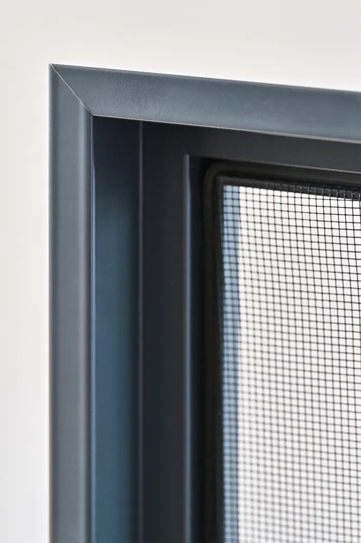 Nahaufnahme eines dunklen Metallfensterrahmens mit einem Insektenschutzgitter, aufgenommen aus einem Winkel, der die Kanten und das Gittermuster des Schutzgitters deutlich macht. Die Befestigung erfolgt mit tesa-Klebeband entlang der Kanten. Der Hintergrund ist einfarbig und unscharf. (This text has been generated by AI)