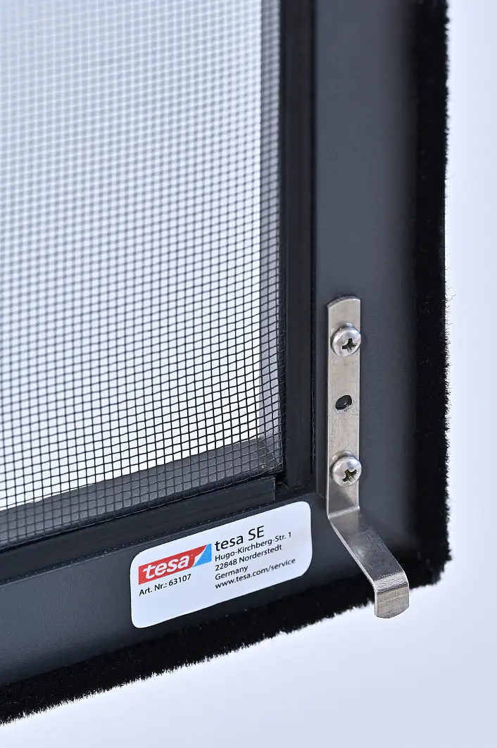 Nahaufnahme eines Fenstergitters mit Metallrahmen, einem Verriegelungsmechanismus und einem Aufkleber mit der Aufschrift „tesa tape“ am unteren Rahmen. Der Hintergrund ist weiß, und das Gittergewebe ist deutlich zu erkennen. (This text has been generated by AI)