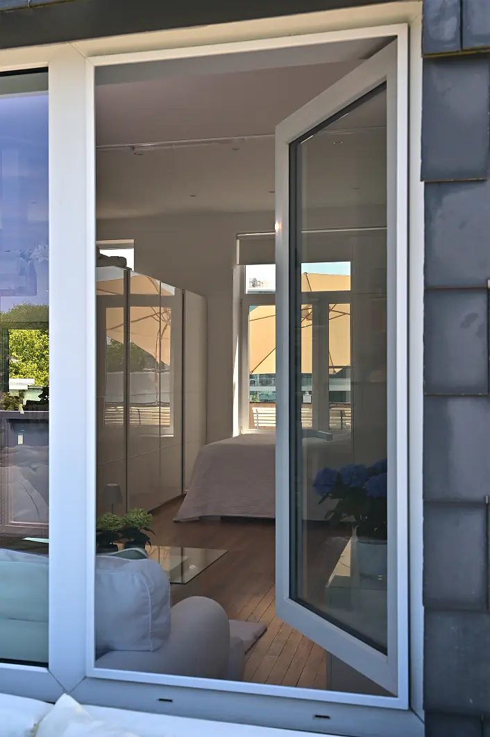 Ein offenes Fenster gibt den Blick auf ein modernes Schlafzimmer frei, in dem ein Bett, weiße Wände, ein Holzboden und blaue Blumen auf einem Tisch zu sehen sind. Sonnenlicht fällt durch weitere Fenster herein und erhellt den Raum. In dieser Szene ist kein tesa-Klebeband zu sehen. (This text has been generated by AI)