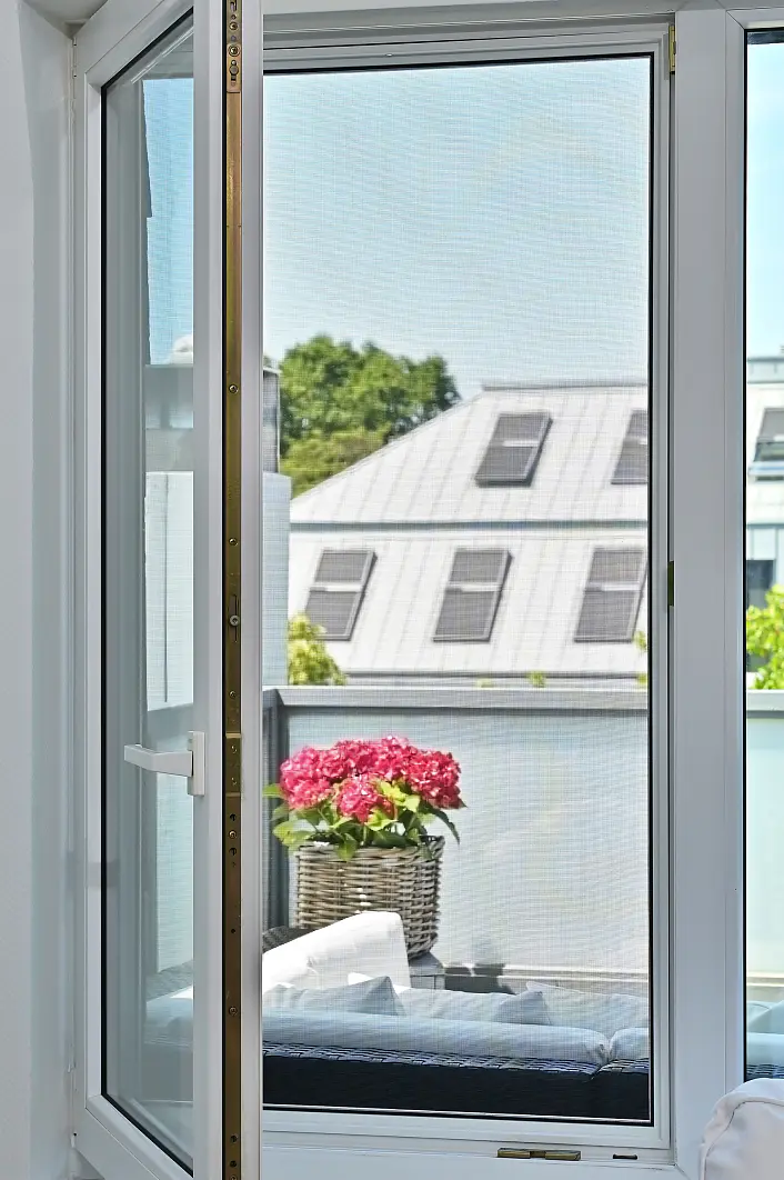 Eine offene Glastür führt auf einen Balkon, auf dem ein Korbstuhl steht, auf dem ein Topf mit leuchtend rosa Blumen steht. Im Hintergrund ist ein Gebäude mit einem grauen Metalldach und zahlreichen Fenstern zu sehen. (This text has been generated by AI)