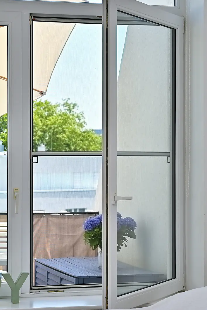 Ein offenes Fenster mit einem Fliegengitter, das mit tesa-Klebeband befestigt ist, bietet den Blick auf einen Balkon. Auf einem blauen Tisch draußen steht eine Topfpflanze mit violetten Blüten. Im Hintergrund sind Bäume und ein Teil eines Gebäudes zu sehen. (This text has been generated by AI)