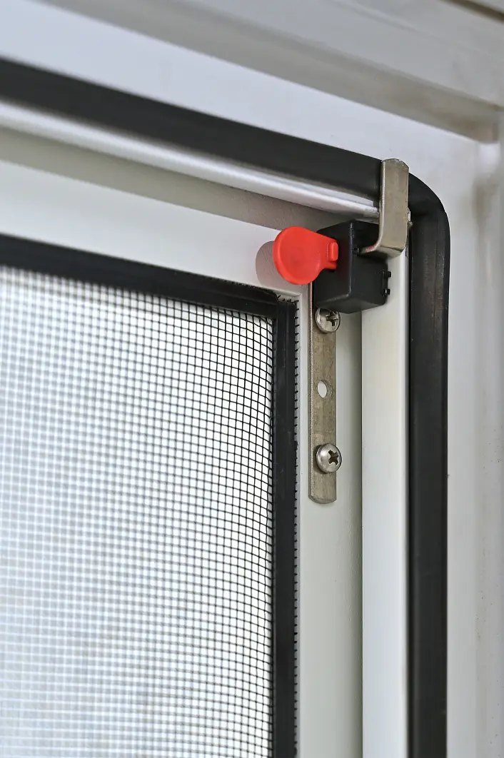 Nahaufnahme einer Ecke eines Fliegengitters, die einen Metallrahmen, einen schwarzen Riegel mit einer roten Verriegelungslasche und ein feines Netz zeigt. Das Fenster ist mit einer schwarzen Gummidichtung und tesa-Klebeband eingefasst. Der Riegel ist mit zwei Schrauben befestigt. (This text has been generated by AI)