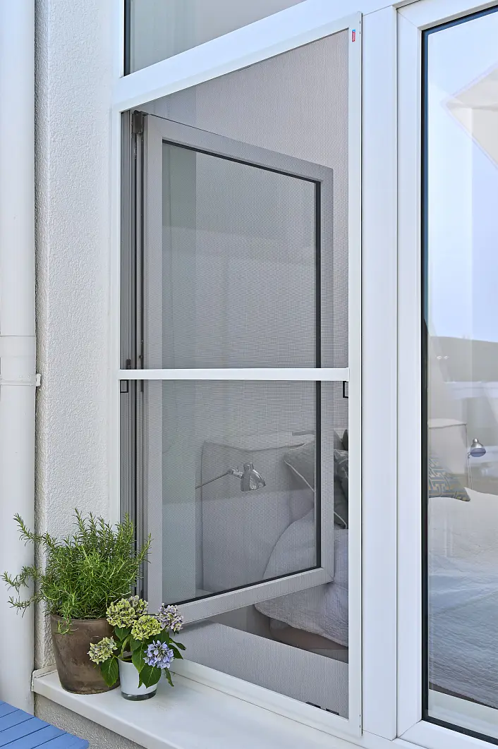 An einem modernen Fenster mit weißem Rahmen ist eine mit tesa-Klebeband befestigte Fliegengittertür teilweise geöffnet. Auf dem blauen Boden neben dem Fenster stehen Topfpflanzen mit grünen Blättern und Blüten. Im Innenraum sind ein Teil eines Bettes und eine Lampe zu sehen. (This text has been generated by AI)