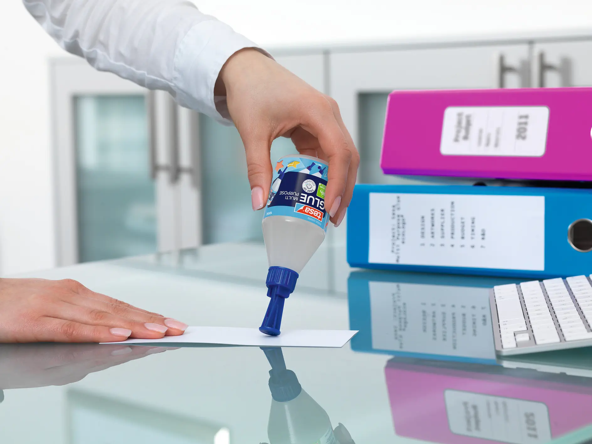 hand applying blue tesa glue from a bottle onto a white sheet of paper on a desk with office folders and keyboard nearby (This text has been generated by AI)