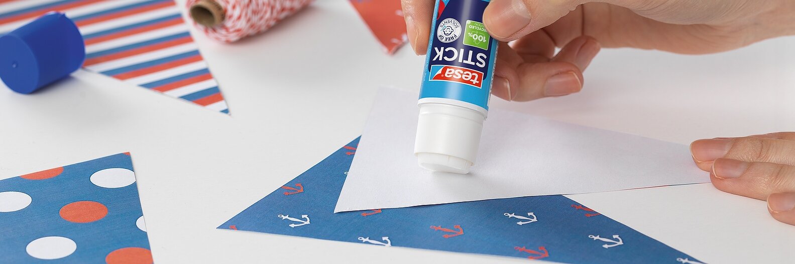 Eine Person trägt Kleber aus einem Klebestift auf ein dreieckiges Stück Papier auf. Auf dem Tisch liegen weitere gemusterte Papierdreiecke sowie Rollen mit tesa-Klebeband und Geschenkpapier.