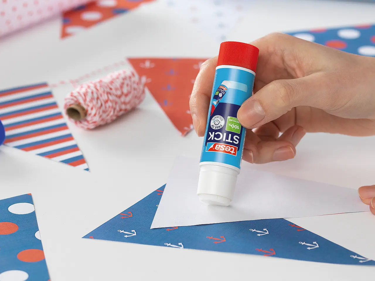 Eine Person trägt Kleber aus einem Klebestift auf ein dreieckiges Stück Papier auf. Auf dem Tisch liegen weitere gemusterte Papierdreiecke sowie Rollen mit tesa-Klebeband und Geschenkpapier.