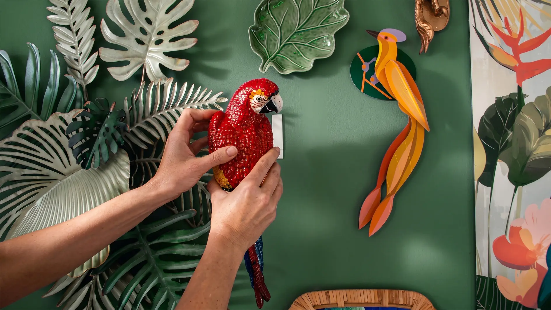 Eine Person befestigt mit tesa-Klebeband eine rote Papageien-Perlenverzierung an einer grünen Wand, die mit Keramikblättern und einem bunten Vogelornament geschmückt ist und von Kunstpflanzen umgeben ist. (This text has been generated by AI)