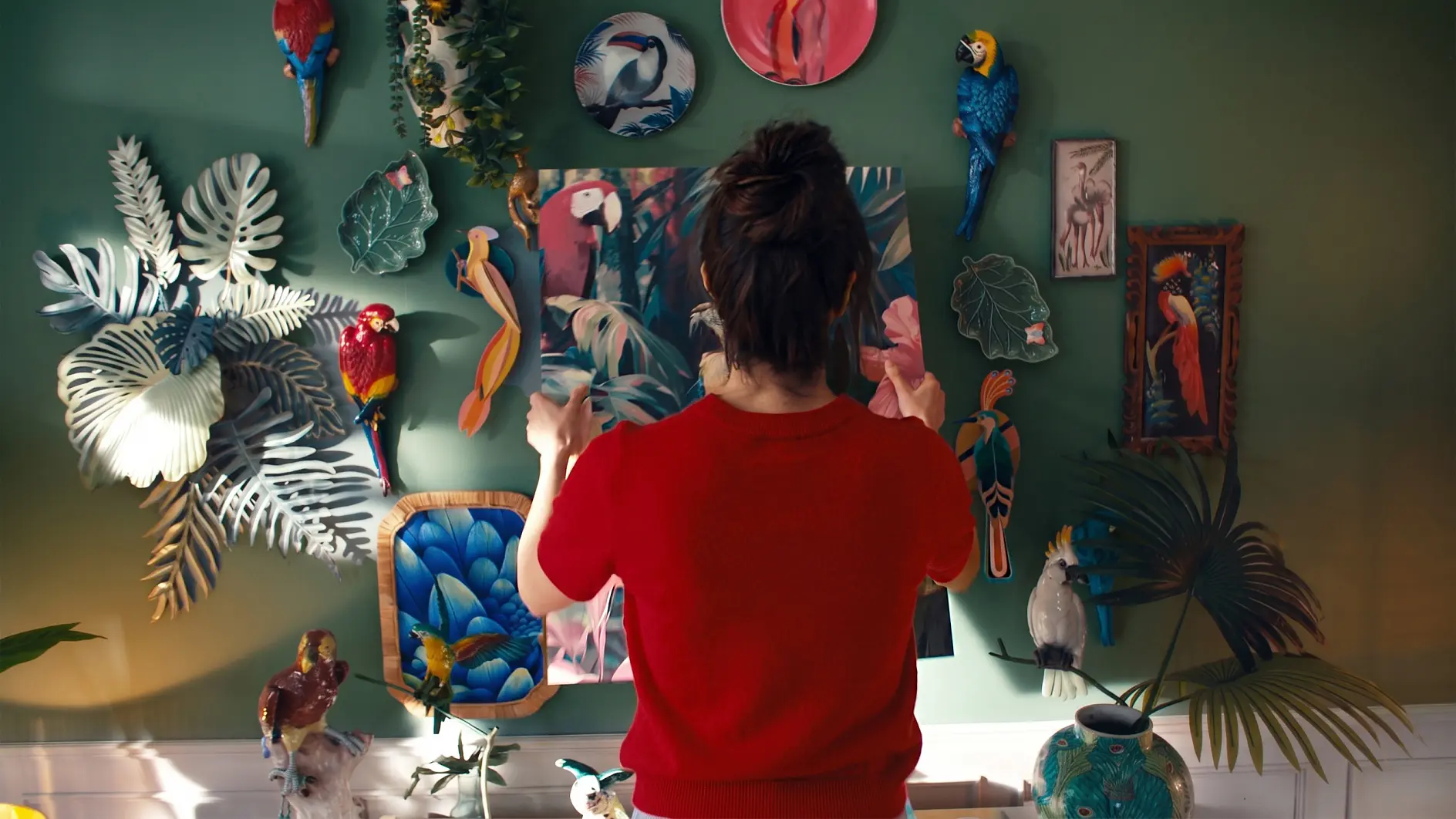 Eine Person mit dunklem Haar, das zu einem Dutt zusammengebunden ist, und einem roten Hemd steht vor einer grünen Wand, die mit bunten Vogelbildern, Pflanzen und Tellern verziert ist, und richtet ein Kunstwerk in der Mitte mit tesa-Klebeband aus oder passt es an.