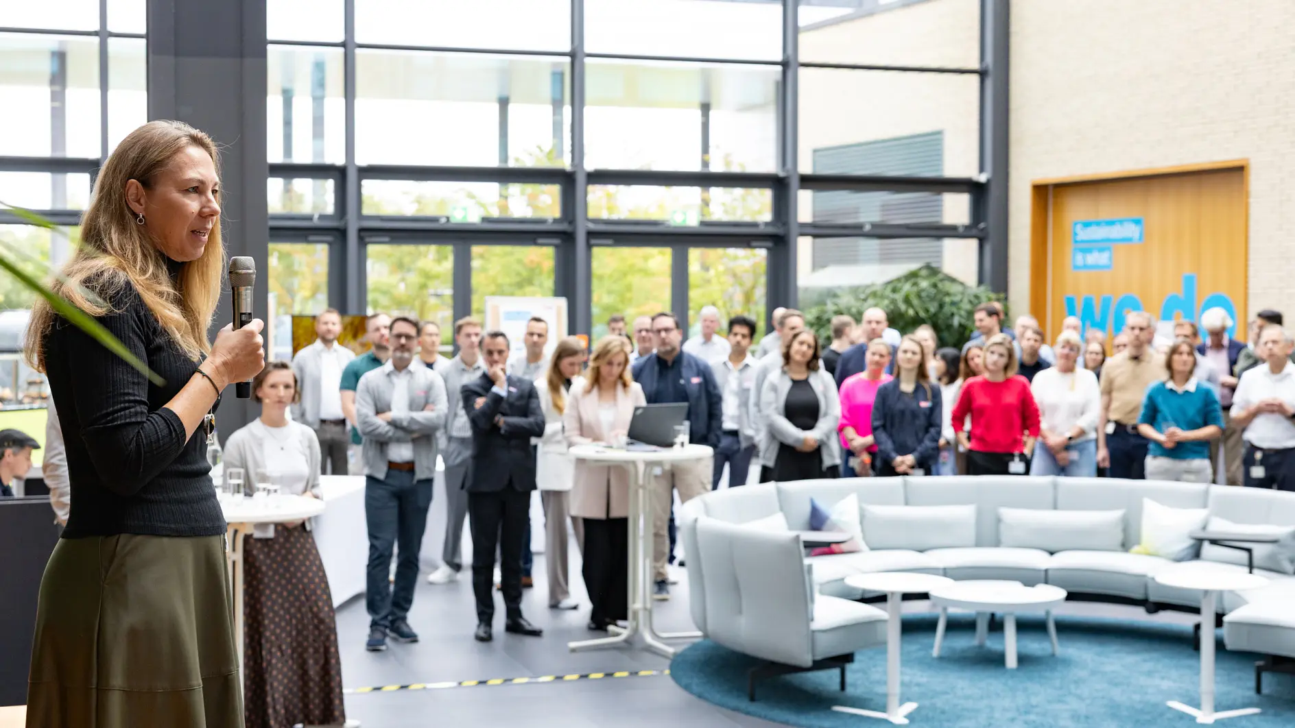 Eine Frau, die in ein Mikrofon spricht, steht vor einem Publikum in einem modernen Büro-Atrium.