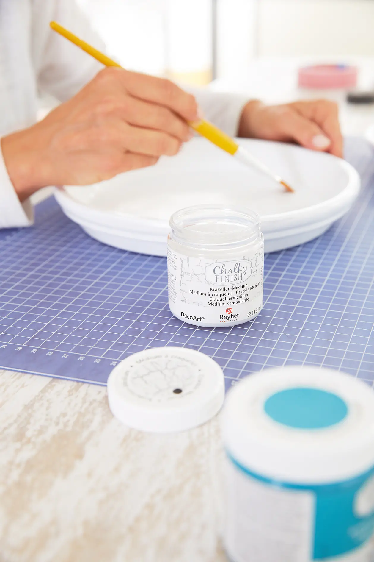 A person is painting a white ceramic plate using a yellow paintbrush. In the foreground, theres a jar labeled Chalky Finish Acrylic Paint on a cutting mat. Another jar with a blue lid is partially visible. (This text has been generated by AI)