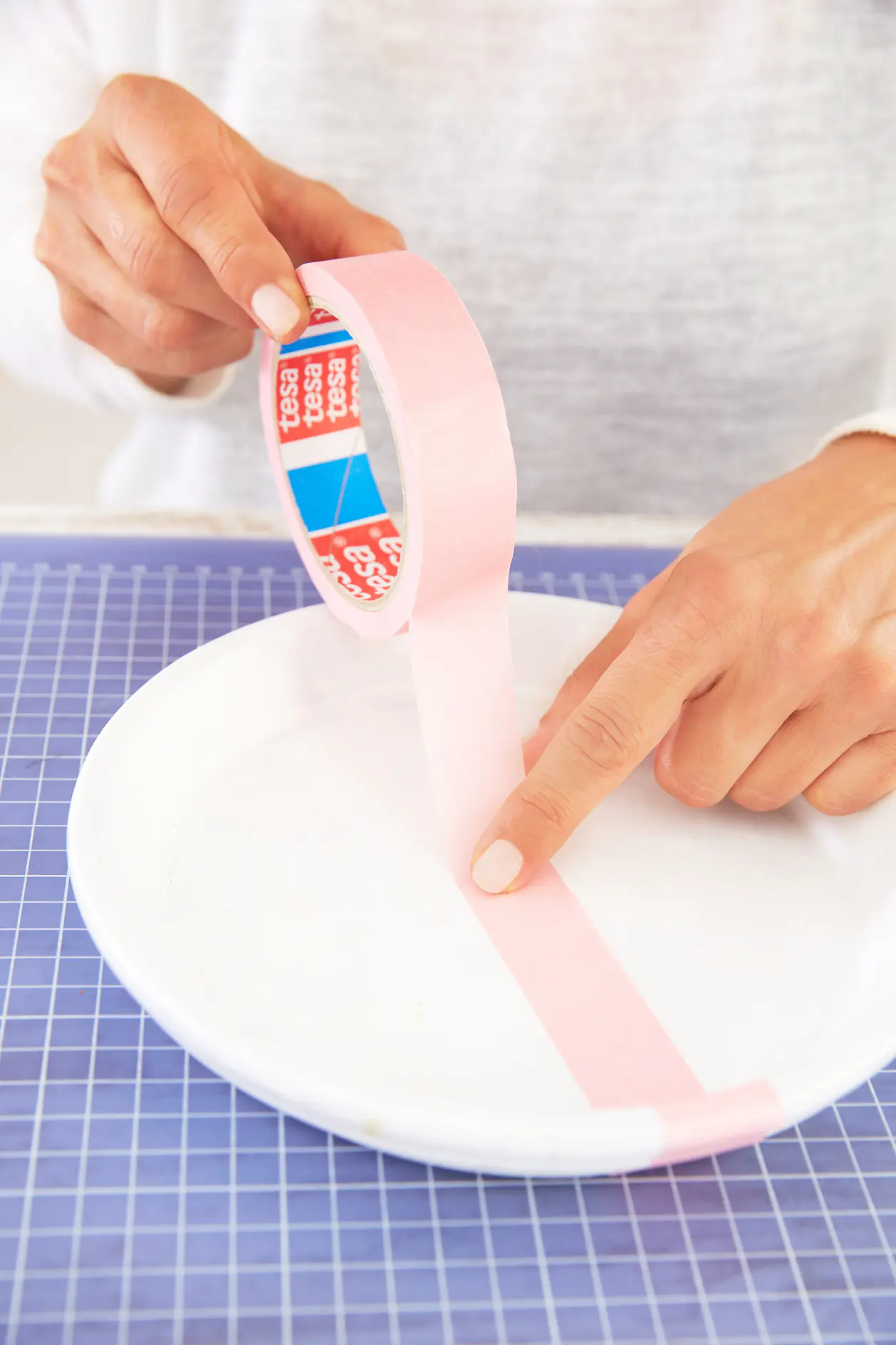 A person is holding a roll of pink tesa tape, applying it to a white plate. The table has a blue grid pattern. The person is wearing a white long-sleeve shirt, and the roll has a red and blue interior. (This text has been generated by AI)