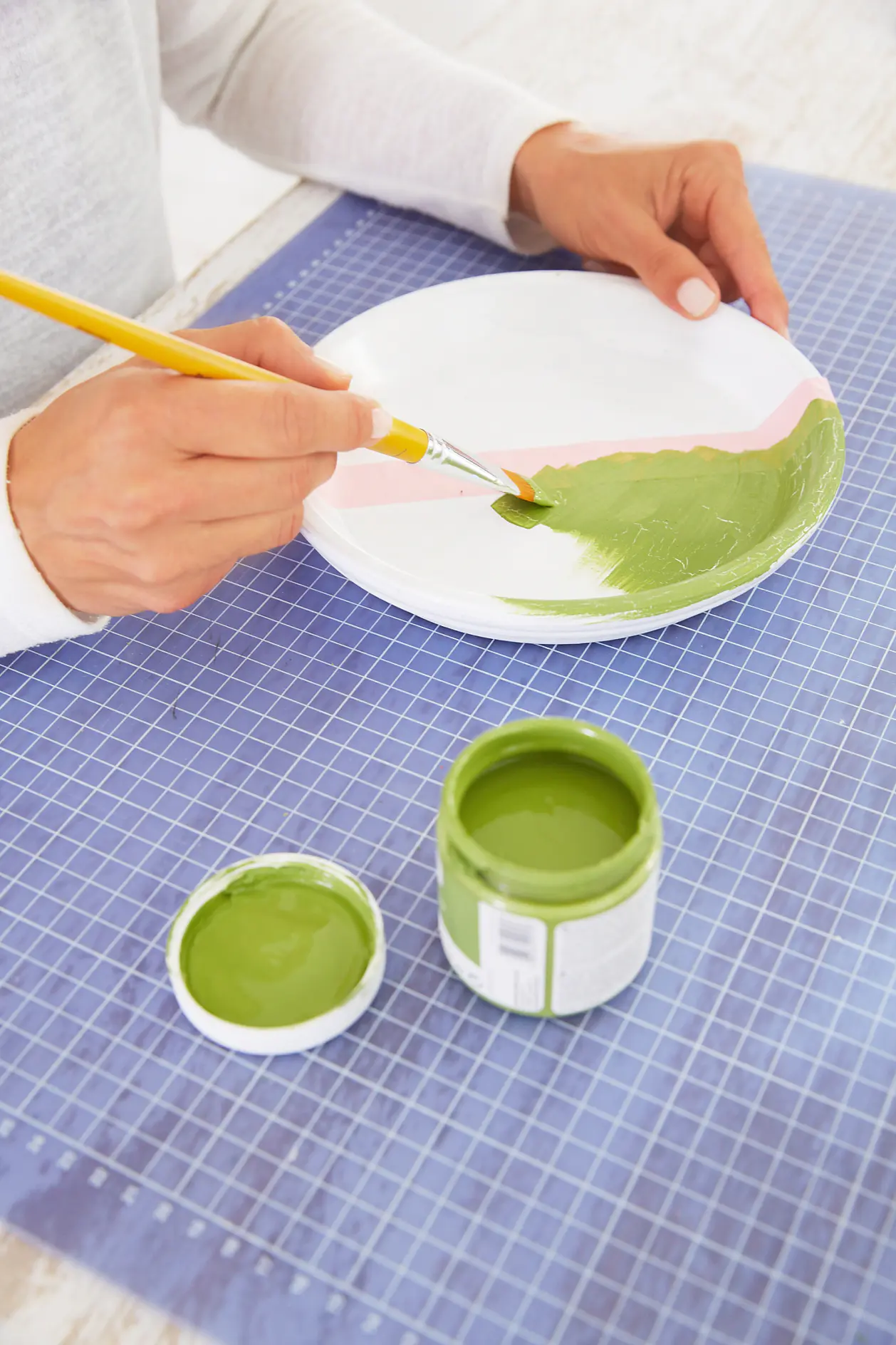 A person is painting a white ceramic plate with green paint using a brush. The plate rests on a blue grid-patterned surface. A small jar of green paint with an open lid is beside the plate. The person is wearing a white long-sleeve shirt. (This text has been generated by AI)
