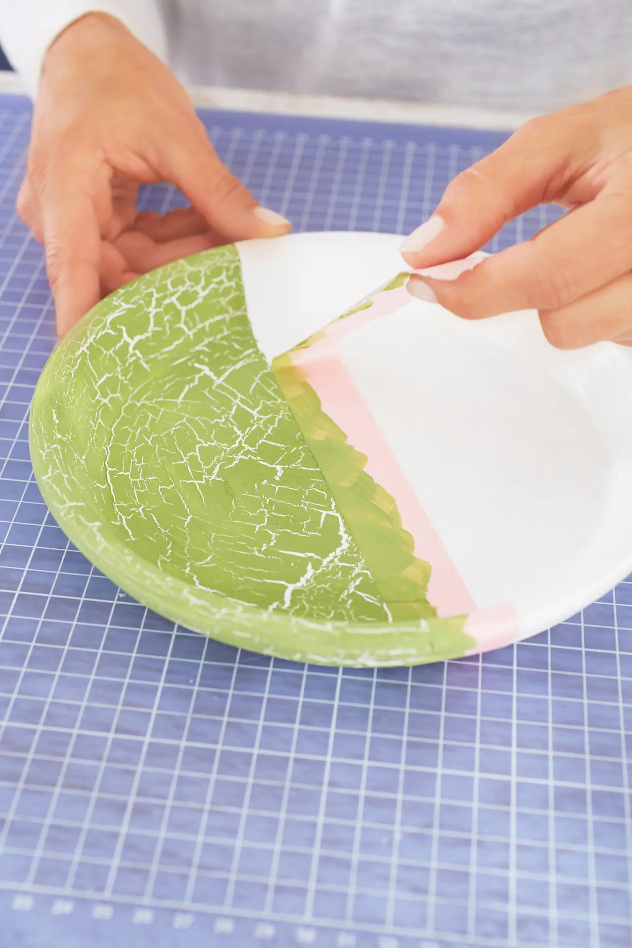 A person is painting a ceramic plate with a brush. The plate is painted with green, pink, and white colors, creating a cracked texture. The plate is on a blue grid-pattern cutting mat secured with tesa tape. (This text has been generated by AI)