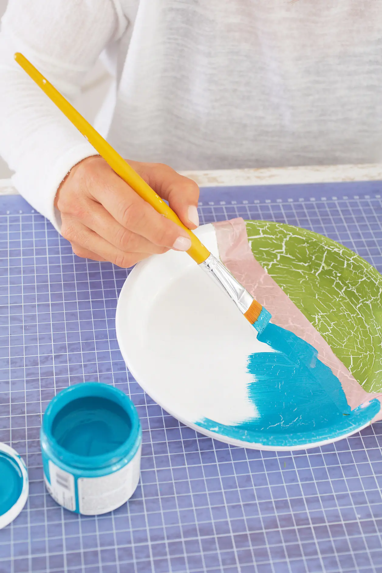 A person is painting a ceramic plate using a flat paintbrush and teal paint. The plate also features sections of green and pink paint with a crackled effect. The person is wearing a white top and is working on a blue grid cutting mat, secured with tesa tape for precision. (This text has been generated by AI)