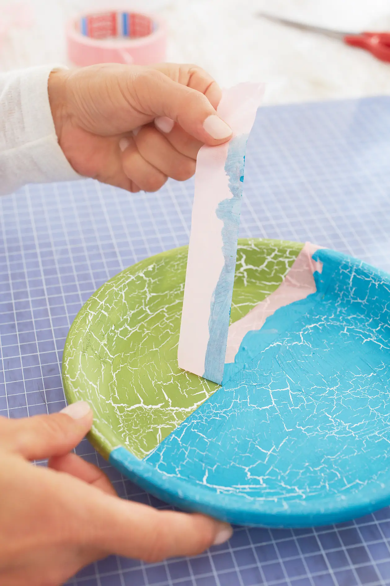 Two hands are peeling off tesa tape from a round plate divided into green and blue sections. The plate has a cracked paint effect. It rests on a blue cutting mat with a grid pattern. Rolls of pink tesa tape and scissors are visible in the background. (This text has been generated by AI)