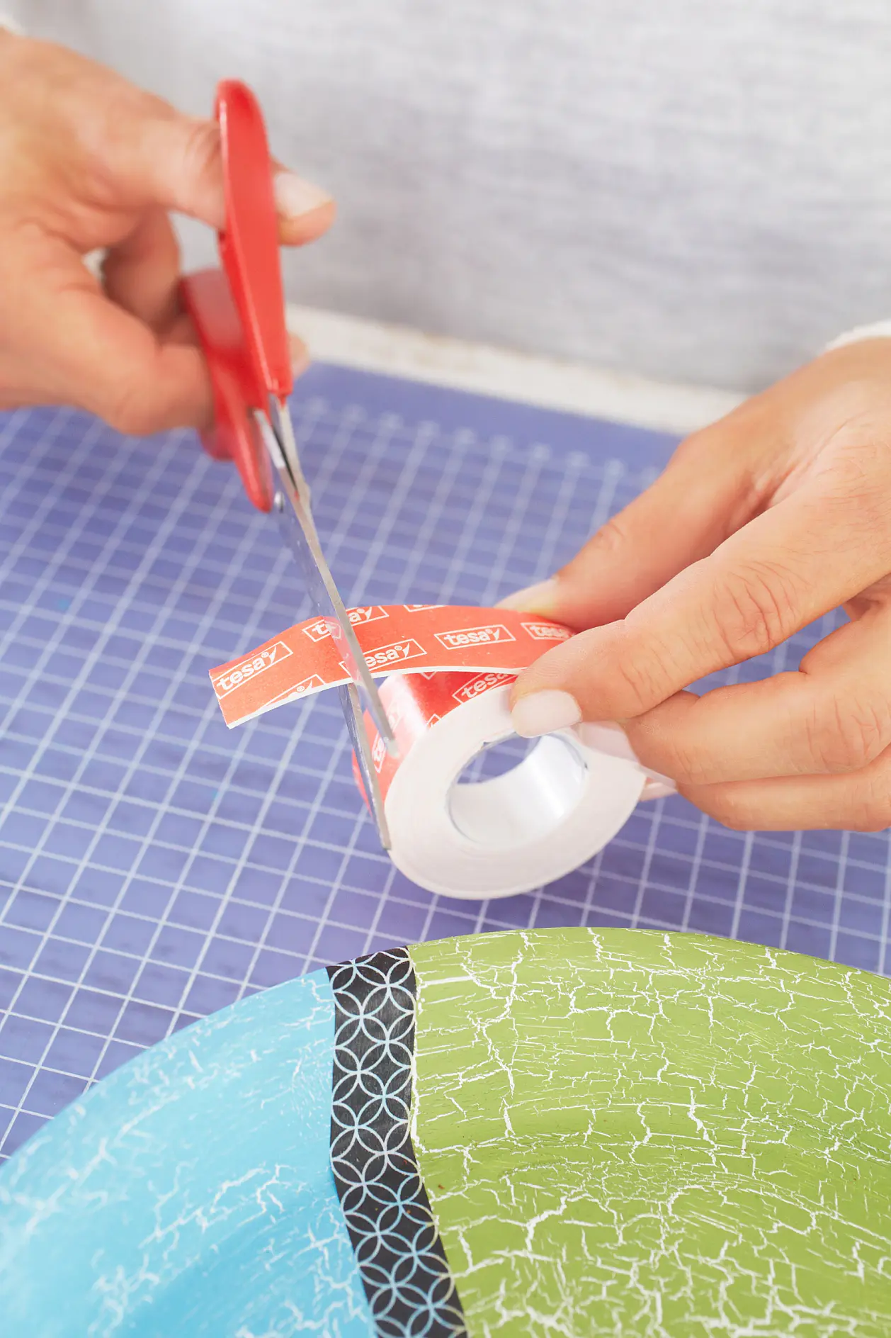 A person uses red scissors to cut a piece of red tesa tape with white text. The tesa tape is being cut over a roll. The background has a blue grid-patterned surface, and a multicolored item with a cracked texture is visible. (This text has been generated by AI)