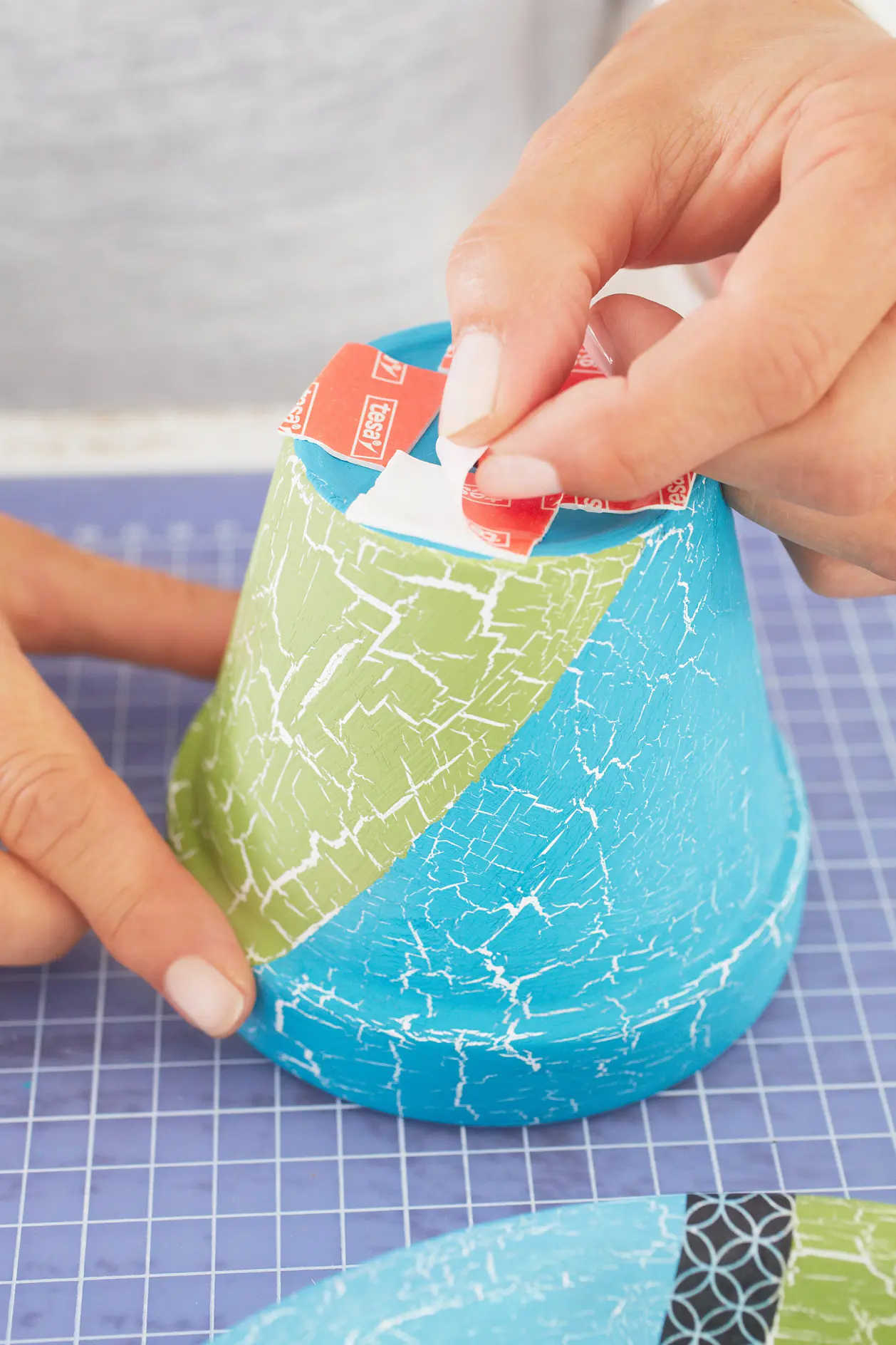 A person is pressing a strip of double-sided adhesive tesa tape onto a small, blue decoupage object. The object has a cracked texture and green parts. The background is a blue grid work surface. (This text has been generated by AI)