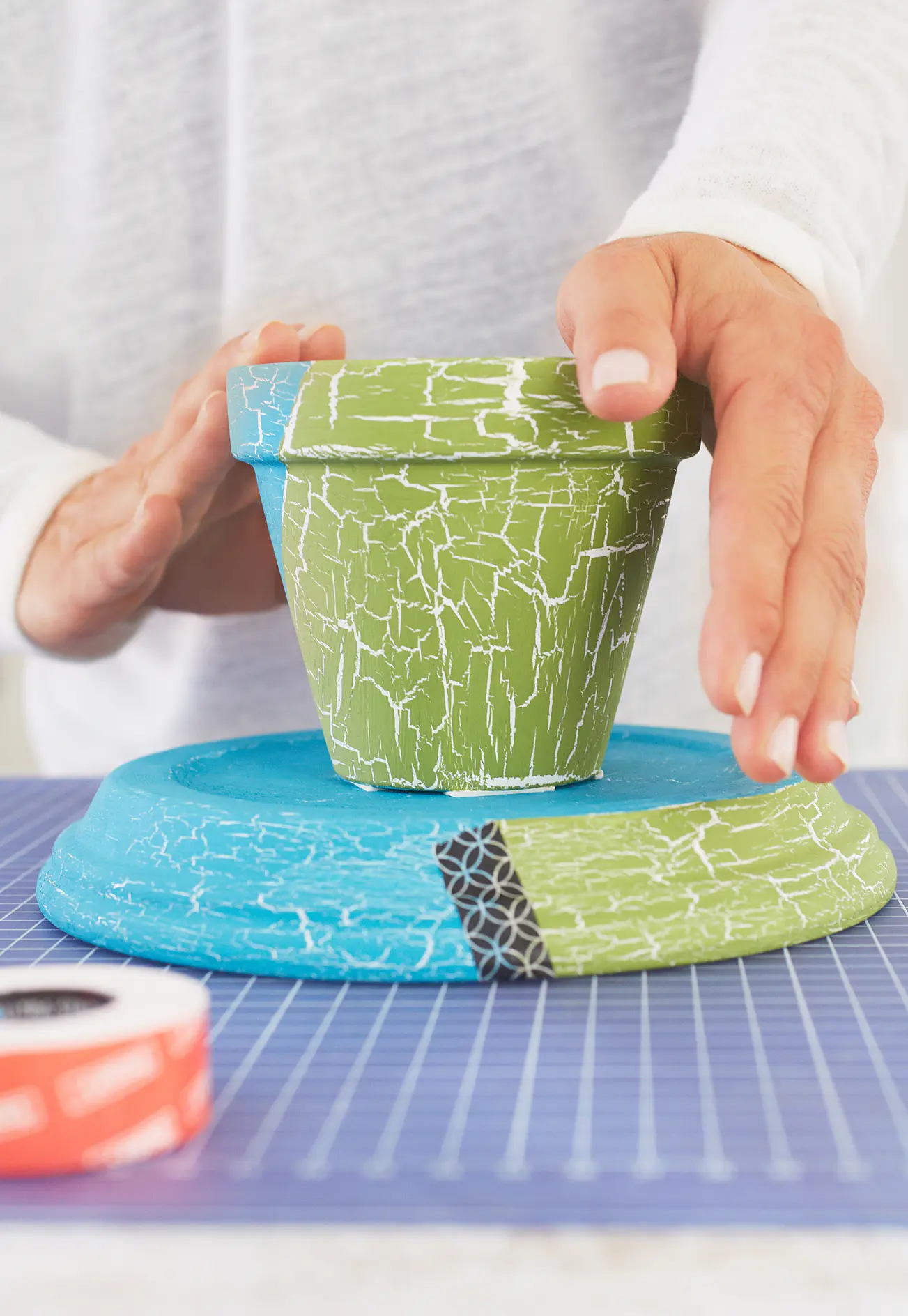 A person is decorating a green clay pot with a crackle effect, placing it on a round, blue platform that also has a crackle pattern. A roll of tesa tape is partially visible in the foreground on a blue grid surface. (This text has been generated by AI)