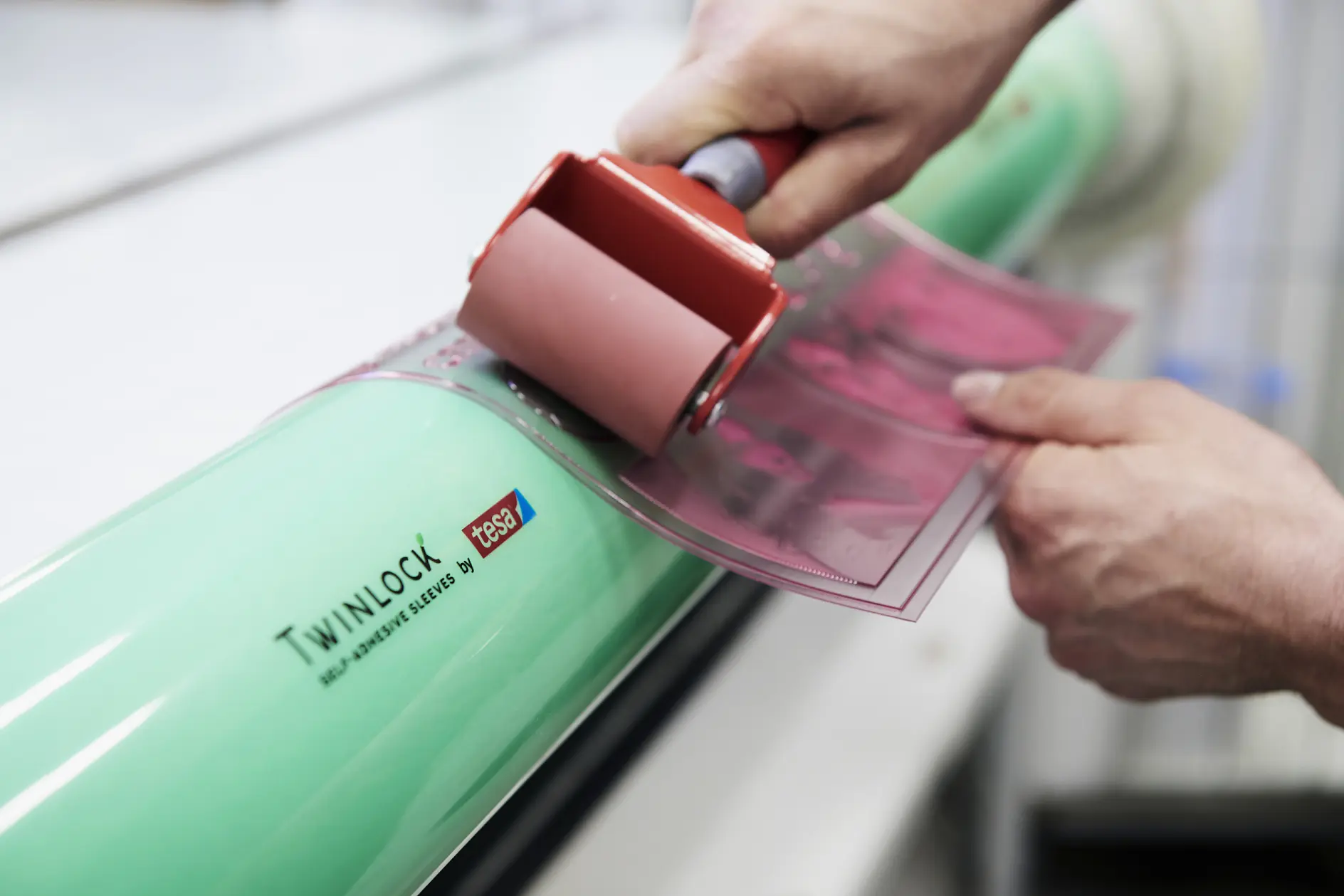 A person uses a red roller to apply a pink film onto a cylindrical green object labeled Twinlock tesa tape. The background is blurred, focusing on the hands and the application process.