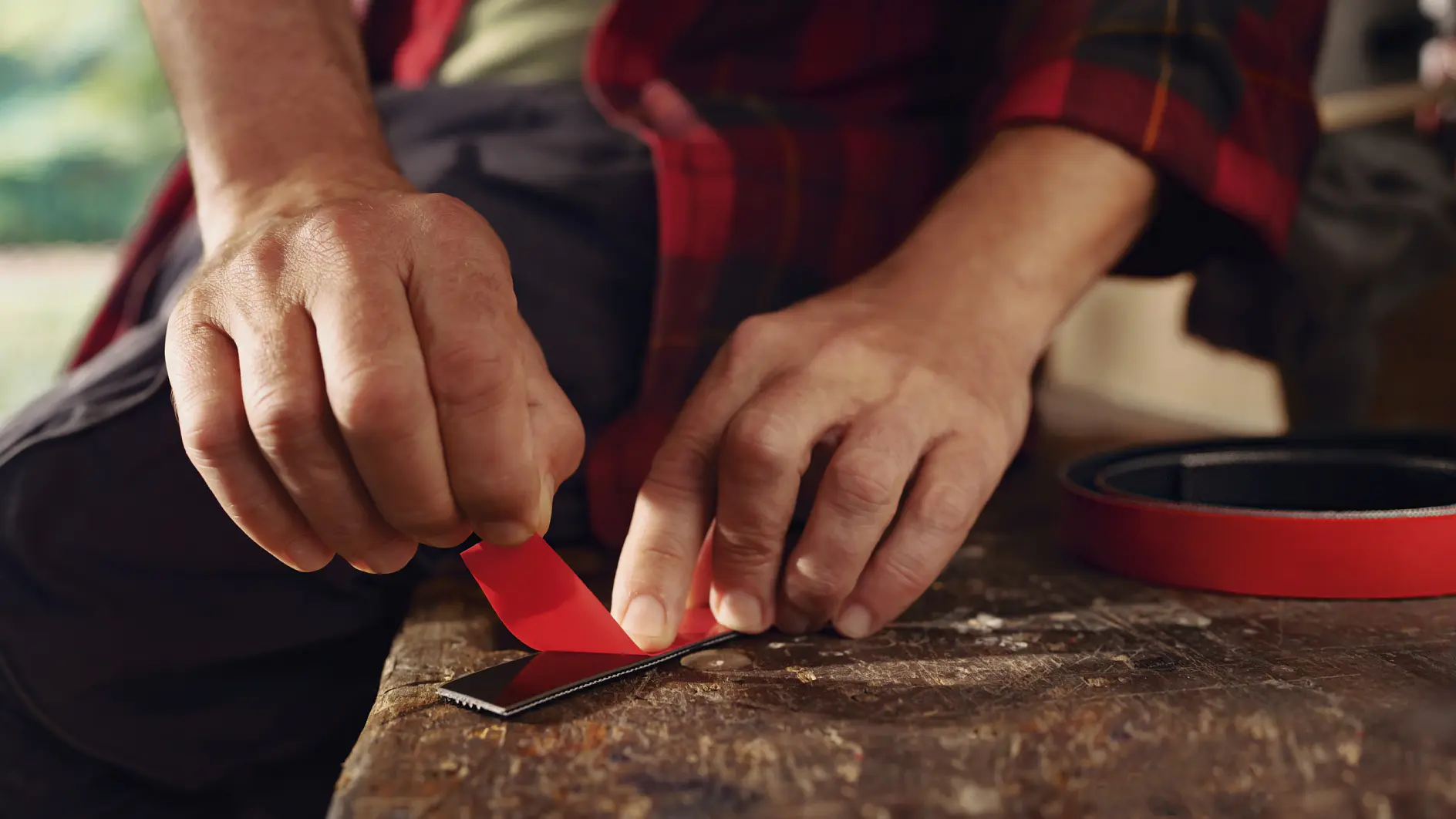 A person in a red and black plaid shirt is cutting red adhesive tesa tape on a wooden surface with a utility knife. A roll of the same tesa tape lies nearby. The persons hands are focused on the task, and the background is slightly blurred. (This text has been generated by AI)