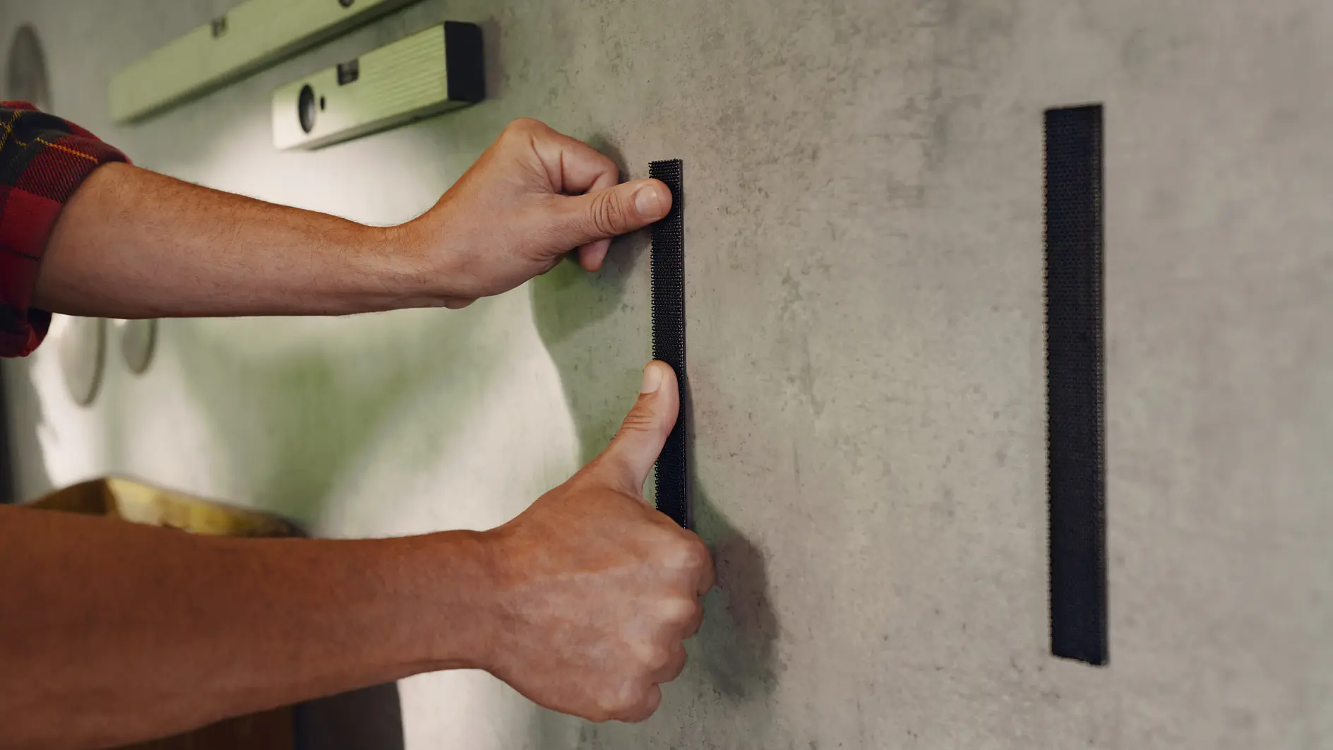 A person is applying two vertical strips of tesa tape to a wall. They press one strip with their thumb while holding the other strip nearby. A level tool is seen in the background on the wall. (This text has been generated by AI)