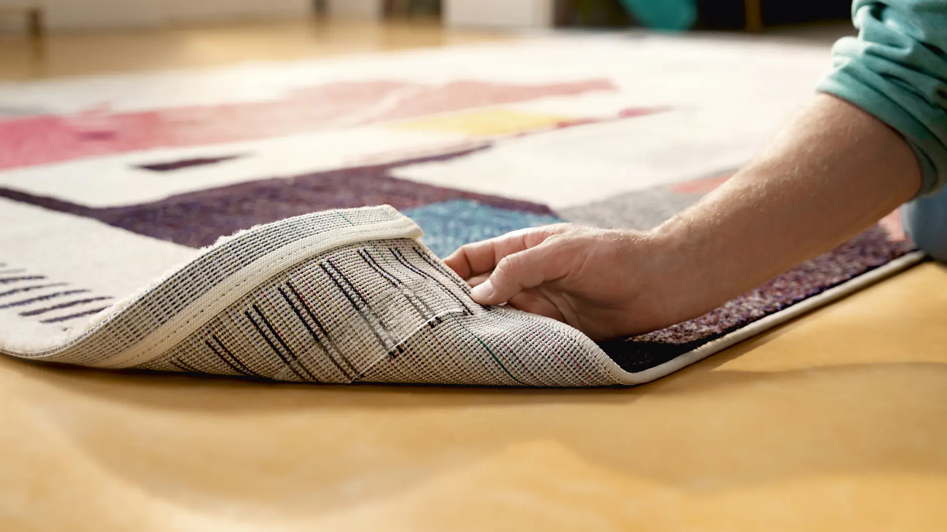 A person lifts the corner of a colorful, geometric-patterned rug, revealing the underside. The rug is placed on a smooth, tan floor using tesa tape. (This text has been generated by AI)