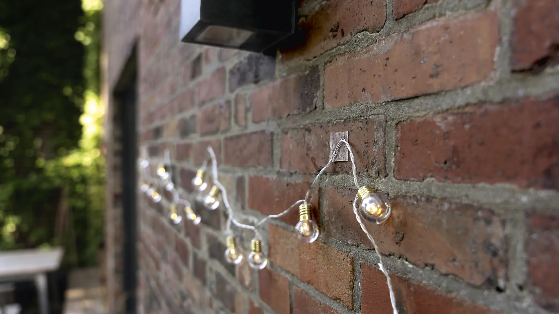 String lights hang along a red brick wall outdoors. The lights are small and spherical, attached to a thin wire. A black rectangular light fixture is mounted above them, and a table is partially visible in the blurred background. (This text has been generated by AI)