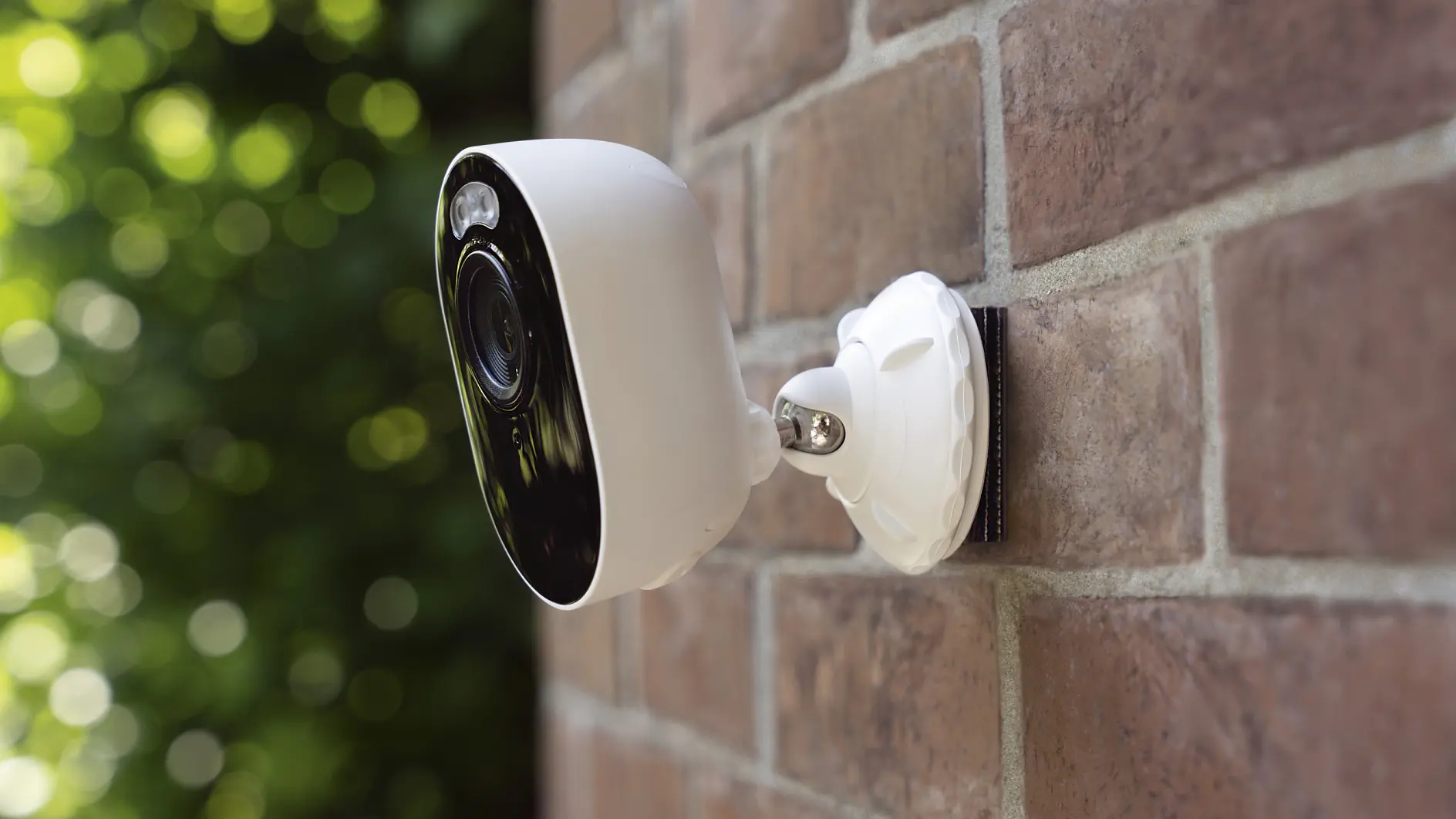 A white security camera mounted on a brick wall outdoors with a blurred background of green foliage. tesa tape is used to secure the camera, which is angled slightly downward, suggesting monitoring of an area below. (This text has been generated by AI)