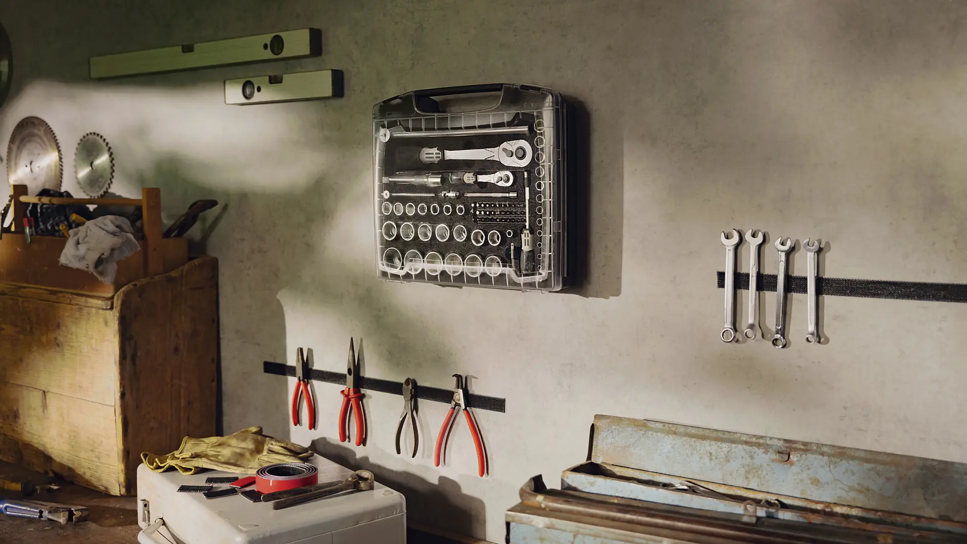 A garage workshop with tools organized on a wall. A clear case with various sockets and wrenches is centrally hung using tesa tape. Below are pliers secured on a magnetic strip fixed with tesa tape. A wooden chest and a blue toolbox are on the workbench, held in place by tesa tape for added stability. A level and saw blades are displayed on the wall, mounted securely using tesa tape. (This text has been generated by AI)