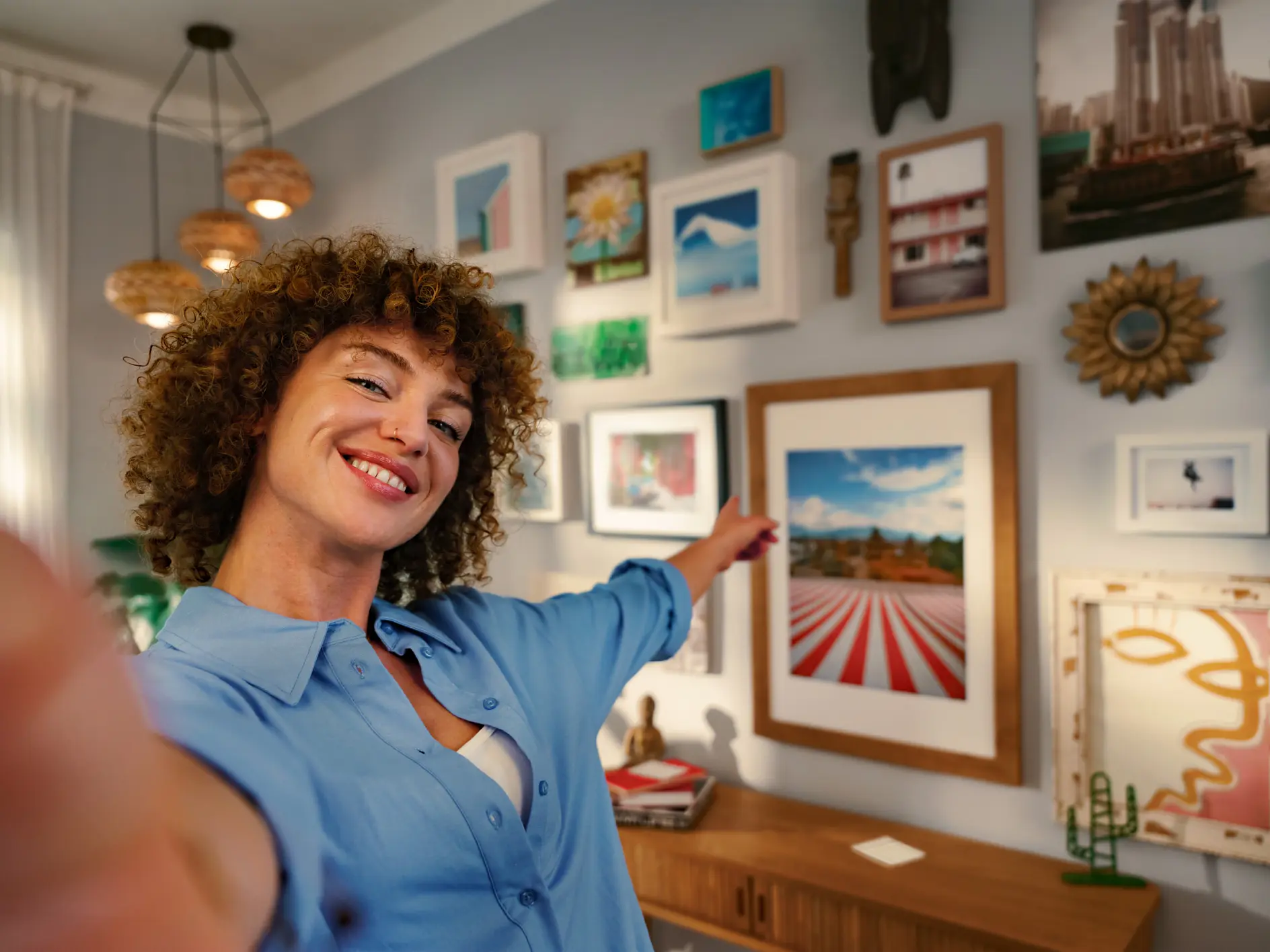 A person with curly hair in a blue shirt takes a selfie while pointing to an art-filled wall. The wall features a variety of framed pictures and art pieces in different sizes and styles. The person is smiling, and the room is warmly lit. (This text has been generated by AI)