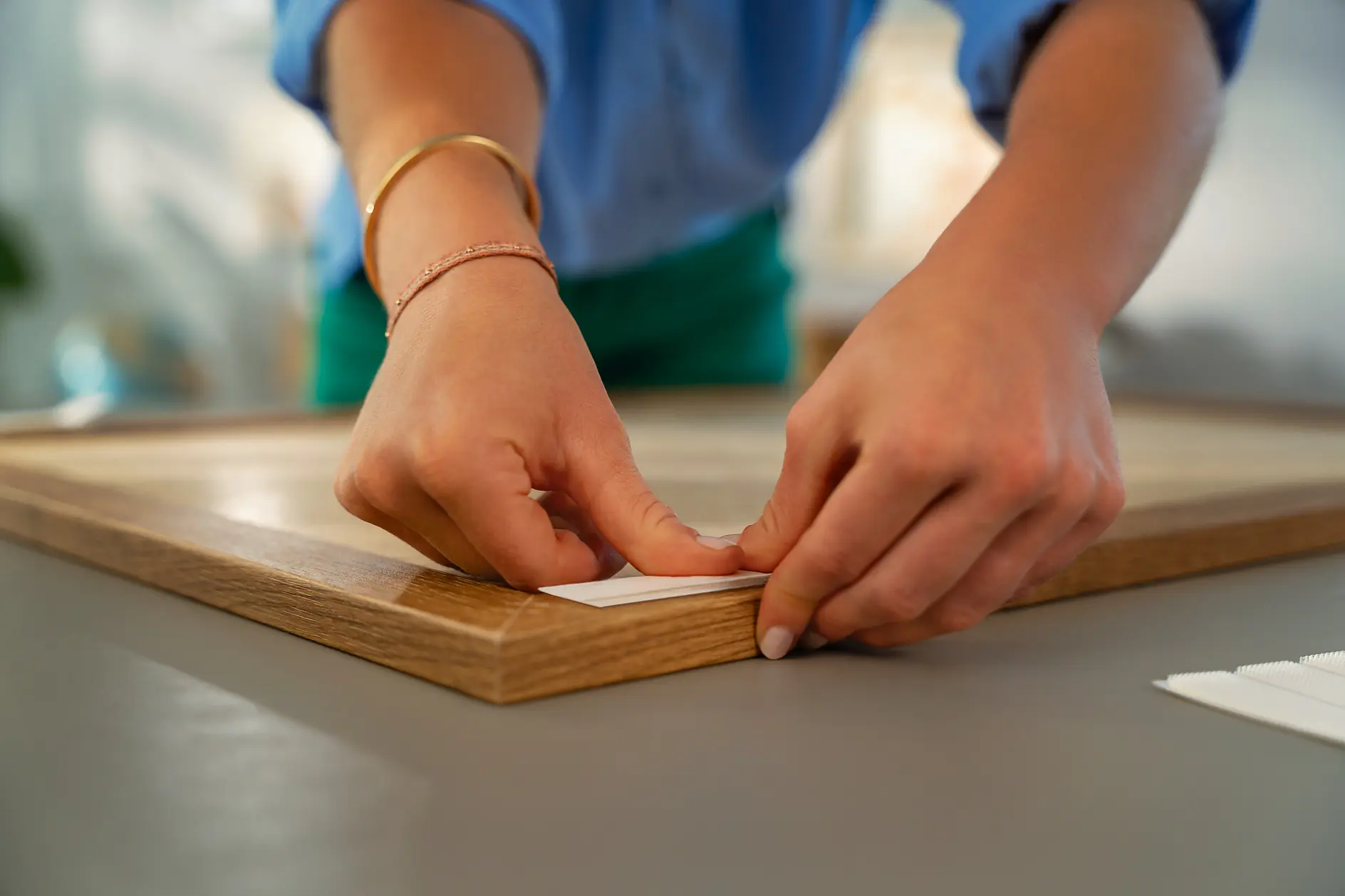 A person wearing a blue shirt and bracelets is attaching tesa tape strips to the back of a wooden frame on a gray surface. (This text has been generated by AI)