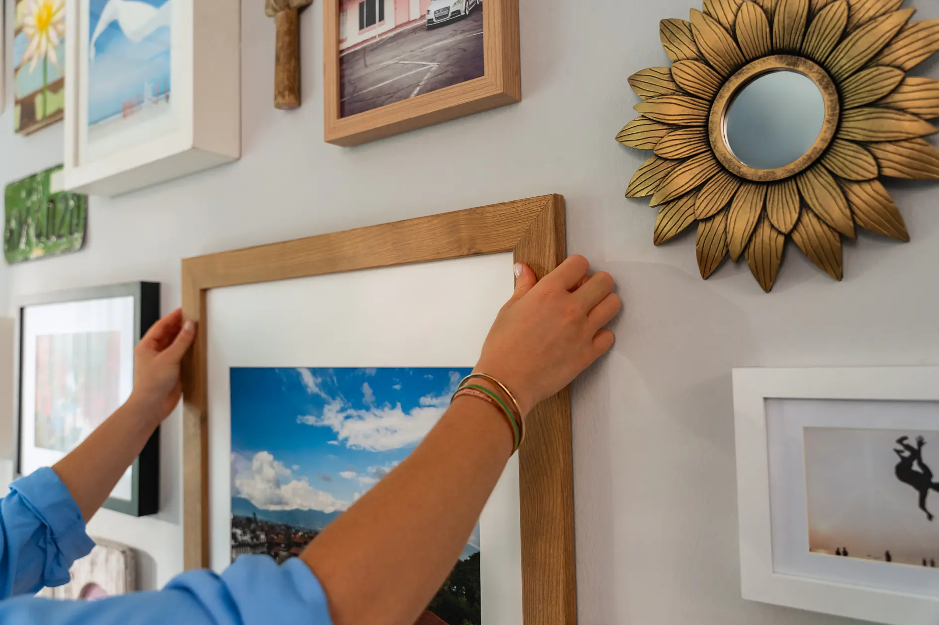 A person wearing a blue shirt is adjusting a framed photograph on a wall. The wall features a gallery of various pictures and art pieces, including a sunflower-shaped mirror. For the adjustments, tesa tape is being used to ensure the picture stays securely in place. The photograph being adjusted shows a landscape with blue skies and clouds. (This text has been generated by AI)