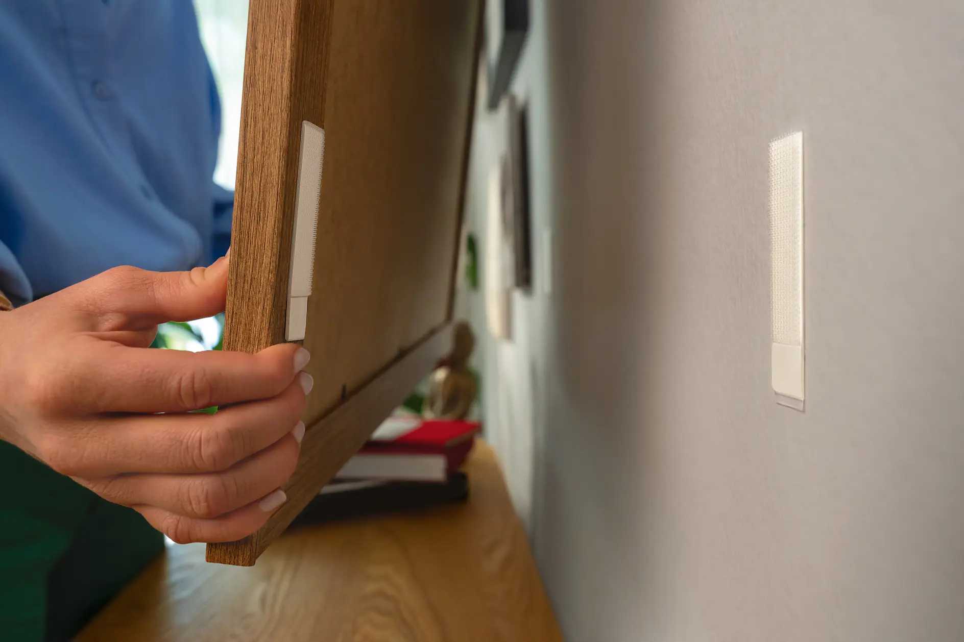 A person in a blue shirt is mounting a wooden frame on a wall using tesa tape adhesive strips. Part of a shelf with items is visible in the background. The frame is positioned near two tesa tape strips on the wall. (This text has been generated by AI)