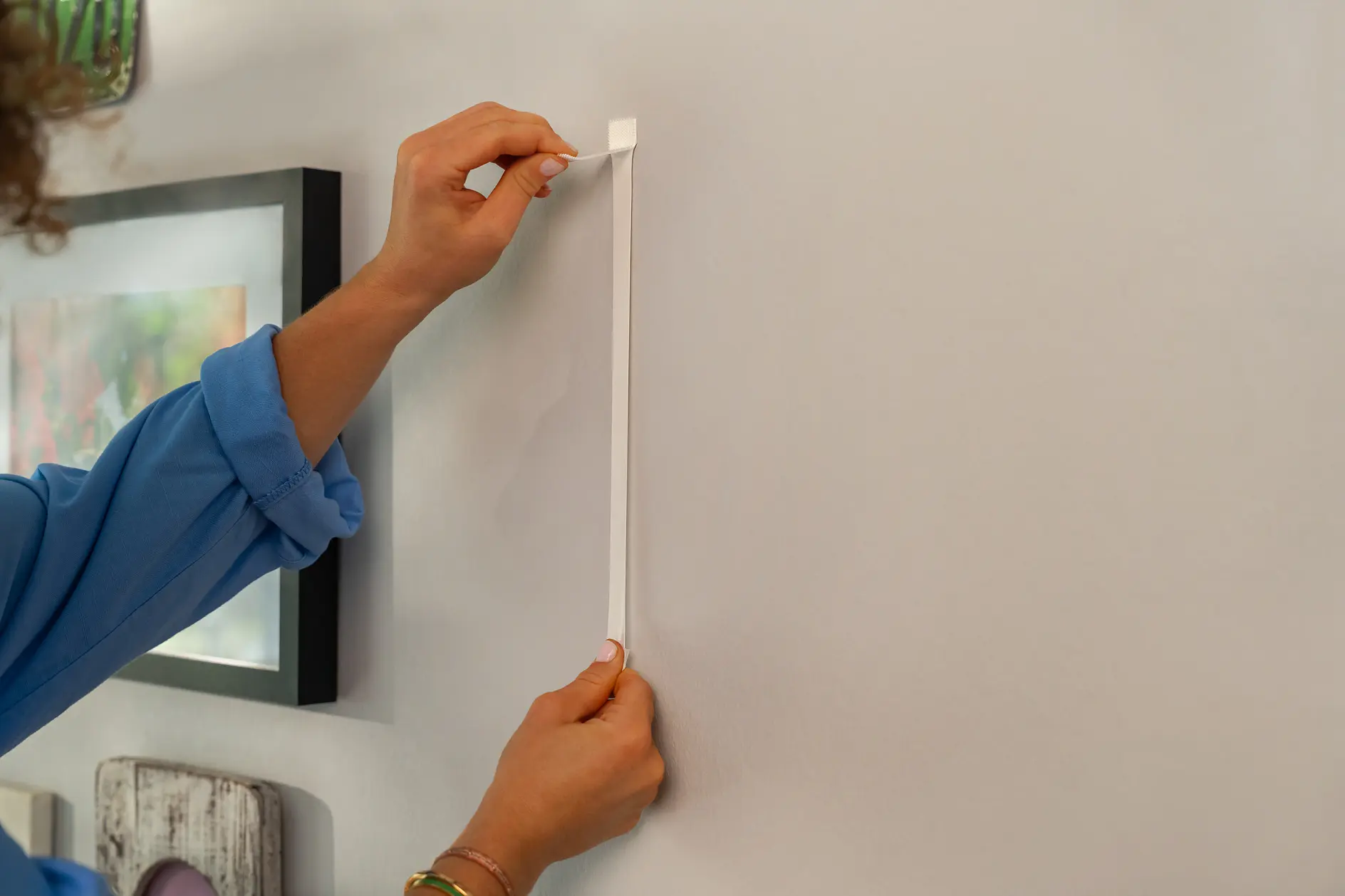 A person in a blue shirt is seen sticking a white tesa tape strip vertically onto a wall. The wall is adorned with multiple framed pictures. The persons hands are positioned to smooth the tesa tape onto the surface. (This text has been generated by AI)