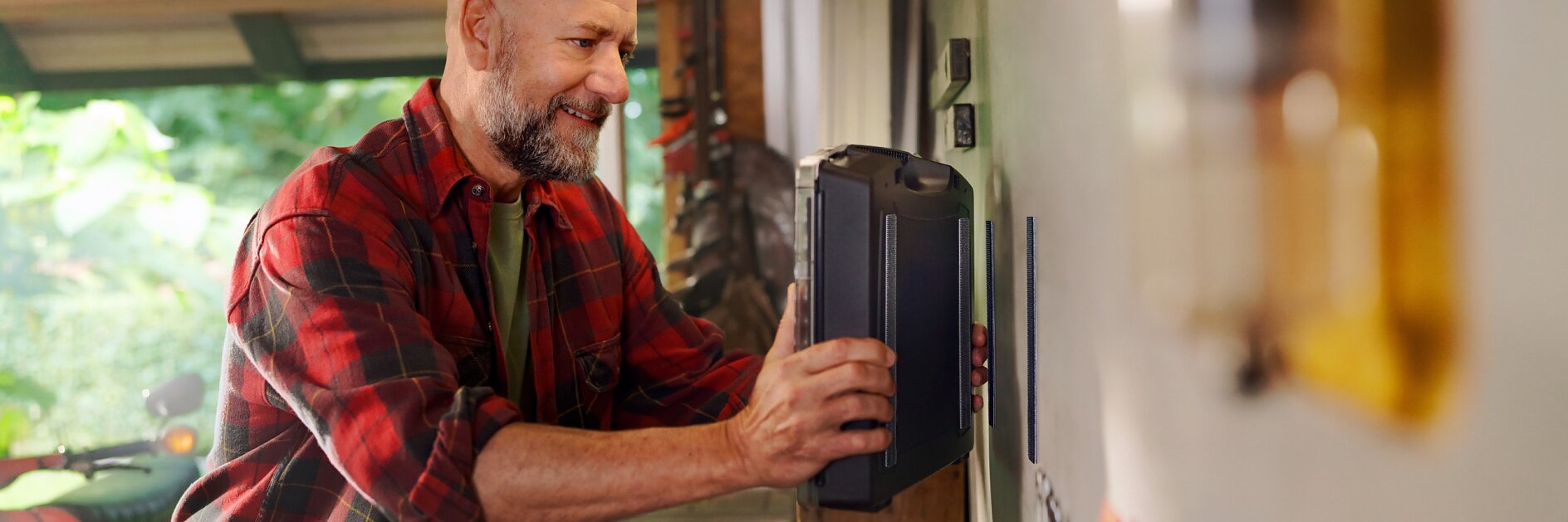 A bald man with a beard, wearing a red plaid shirt, is placing a toolbox on a shelf in his workshop. He smiles as he organizes tools around him. The workshop is brightly lit with various tools and equipment visible in the background, along with tesa tape prominently displayed among them.